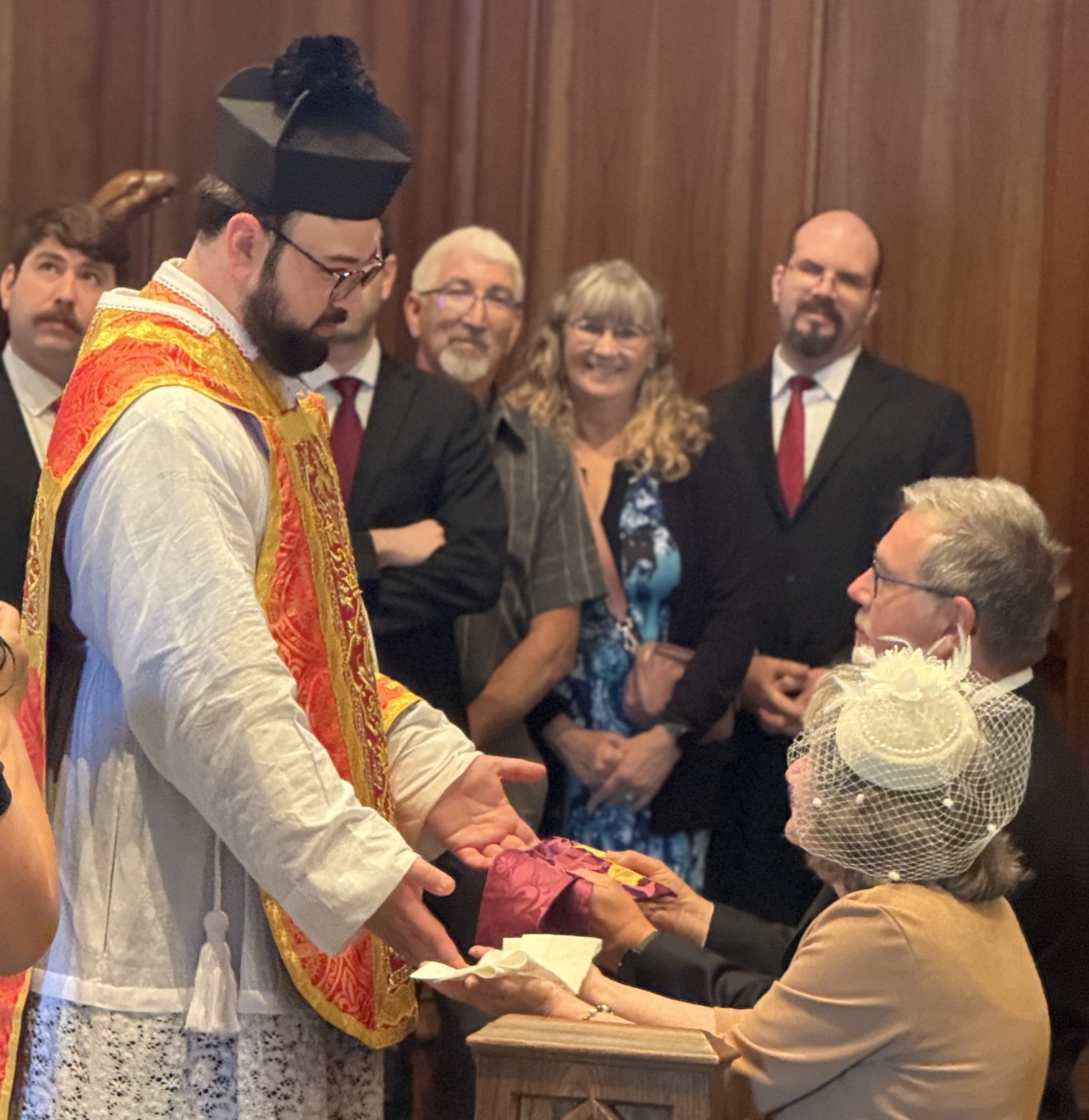Fr. Andrew Yaremko, in vestments and biretta, hands maniturgium to mother and maniple (seemingly) to father as both kneel